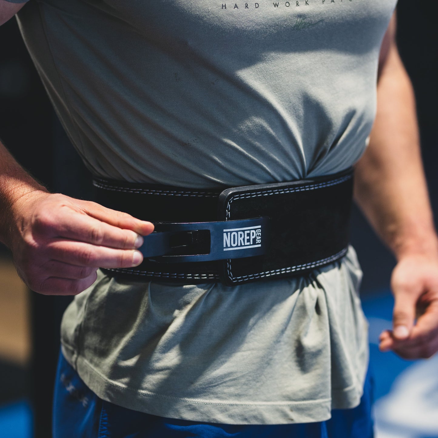 Person wearing a gray t-shirt with visible branding and blue pants, focusing on a black belt with a brand logo.