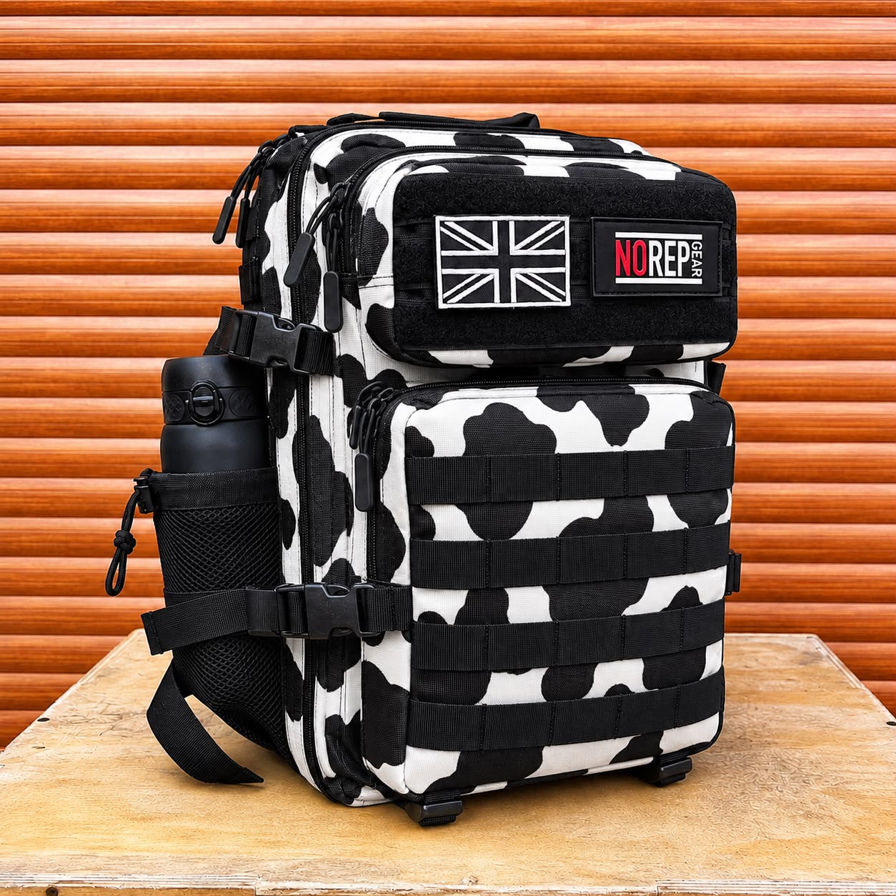 A tactical-style backpack with a black and white cow-print pattern, black straps, and a side water bottle holder. 