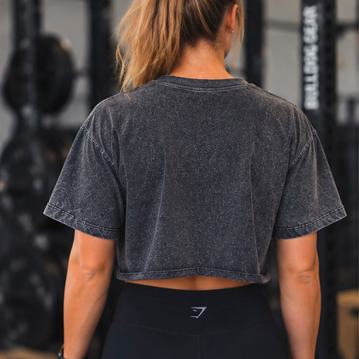 A woman viewed from behind wearing a washed charcoal gray cropped t-shirt and black leggings in a gym setting.