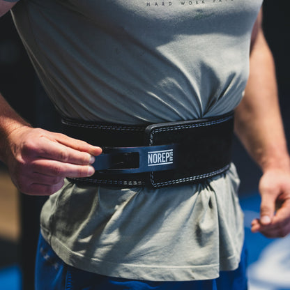 Person wearing a gray t-shirt with visible branding and blue pants, focusing on a black belt with a brand logo.