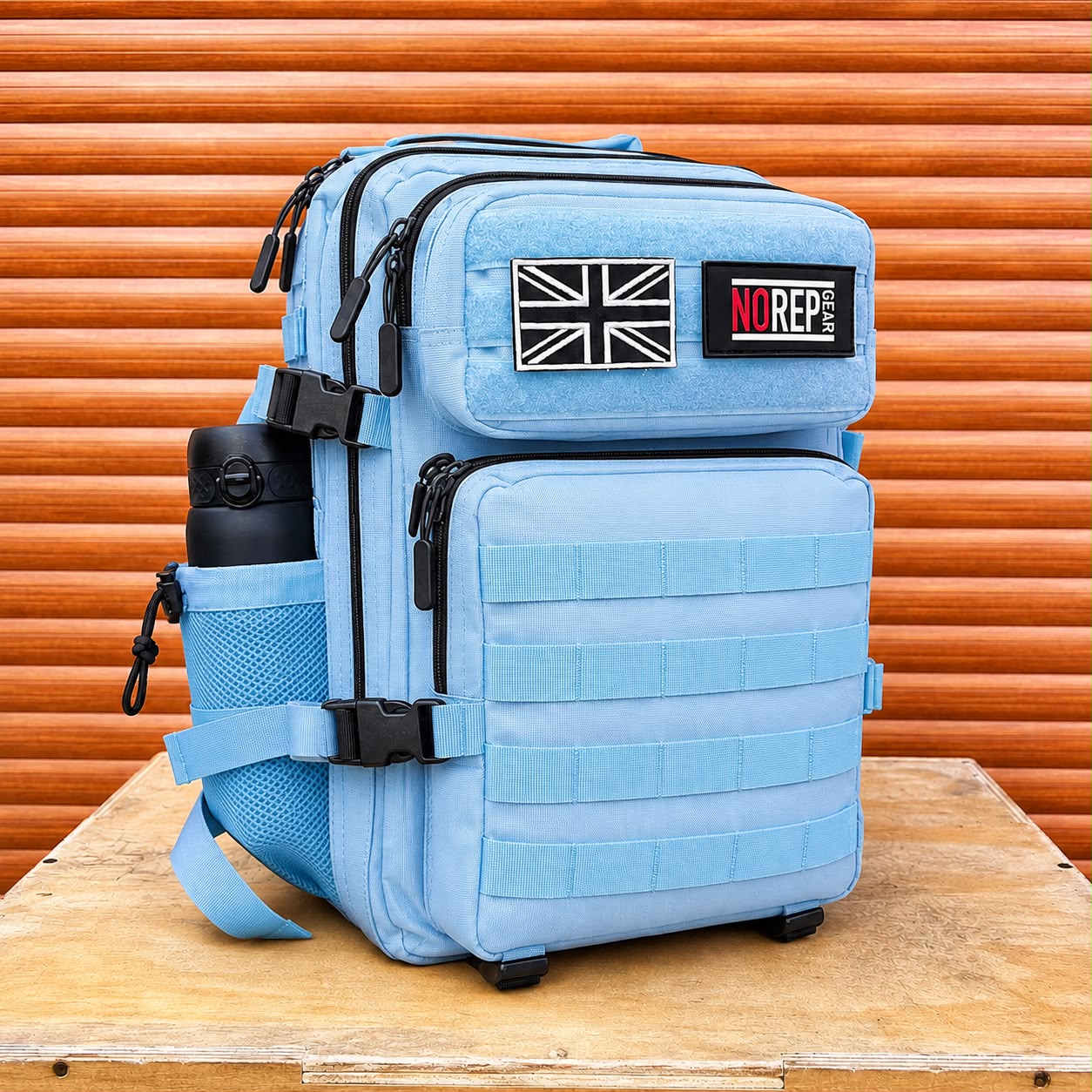 A light blue tactical backpack with black zippers, webbing, and a side mesh bottle holder, standing on a wooden surface in front of an orange corrugated metal background. The backpack features a Union Jack patch and a black 'NOREP' brand patch on the front.